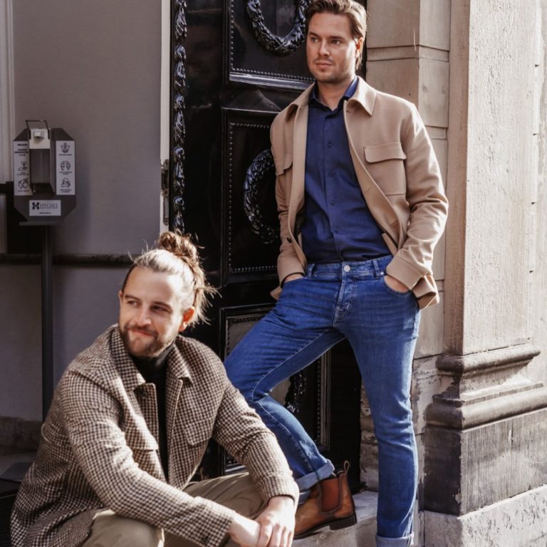 Twee mannen dragen stijlvolle, casual outfits in een stadsomgeving. De een staat bij een deuropening in een beige jasje, blauw overhemd en spijkerbroek; de ander zit in een geruite jas en olijfgroene broek, met een knotje en een baard. Het zonlicht verlicht de scène en benadrukt hun ontspannen poses. | Hoes Tailors Alkmaar