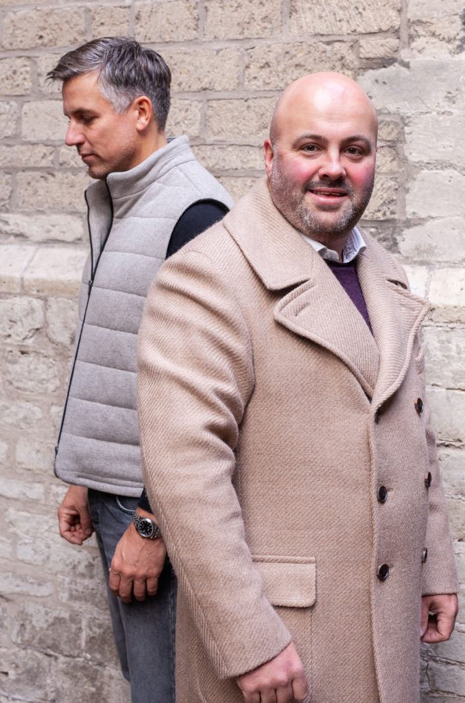 Twee mannen staan rug aan rug voor een stenen muur. De man links heeft grijs haar en draagt een grijs gewatteerd vest en een zwart shirt, en kijkt naar beneden. De man rechts is kaal, heeft een baard, draagt een bruine jas en lacht naar de camera. | Hoes Tailors Alkmaar