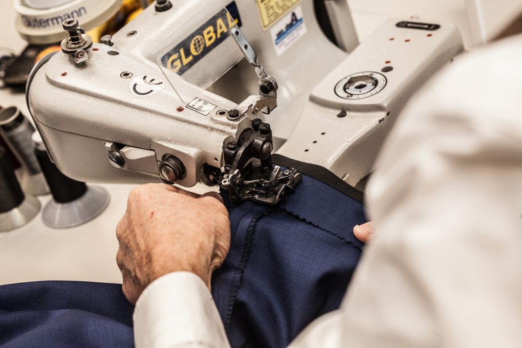Een close-up van de handen van een kleermaker die een naaimachine gebruikt om donkerblauwe stof te naaien. De garenklossen en bedieningselementen van de machine zijn zichtbaar, en de persoon draagt een wit overhemd en concentreert zich op het precieze naaiwerk langs de rand van de stof. | Hoes Tailors Alkmaar