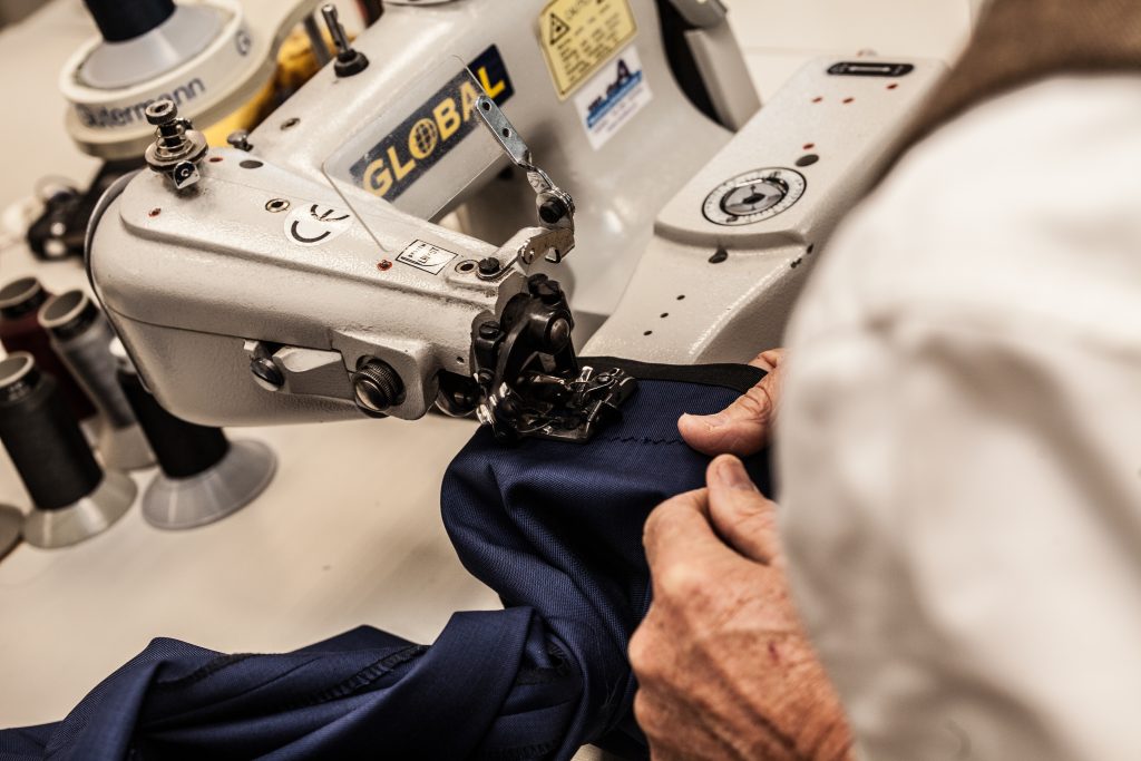 Close-up van een persoon die donkerblauwe stof naait met een industriële naaimachine in een werkplaats. Alleen de handen en een deel van de torso zijn zichtbaar, waardoor de nadruk ligt op het precisiewerk. Op de achtergrond zijn verschillende klosjes garen te zien. | Hoes Tailors Alkmaar