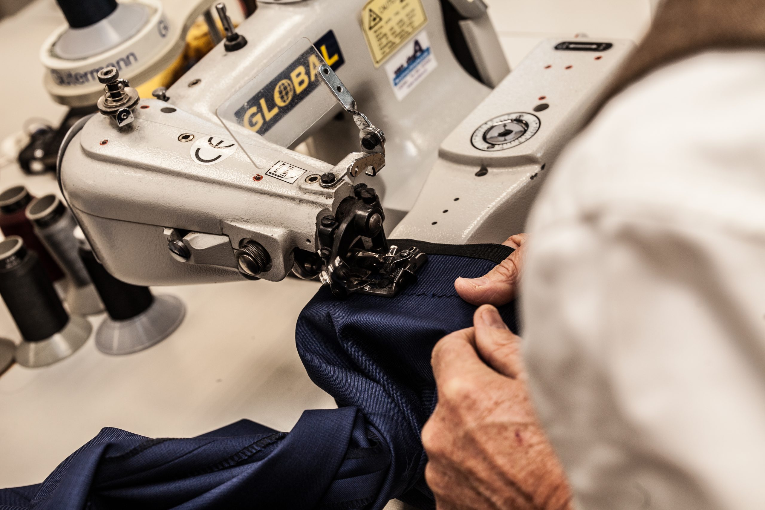 Close-up van een persoon die donkerblauwe stof naait met een industriële naaimachine in een werkplaats. Alleen de handen en een deel van de torso zijn zichtbaar, waardoor de nadruk ligt op het precisiewerk. Op de achtergrond zijn verschillende klosjes garen te zien. | Hoes Tailors Alkmaar