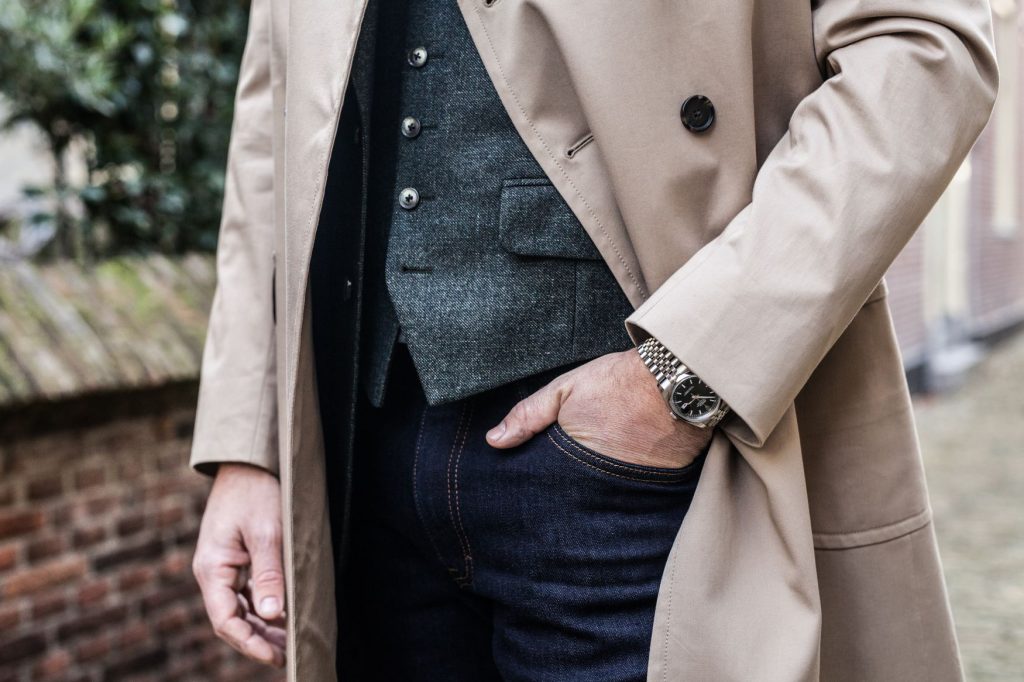 Een persoon draagt een beige trenchcoat over een grijs vest met knoopjes en een donkere spijkerbroek, met één hand in de zak. Om de pols draagt hij een zilveren horloge met een metalen band. De achtergrond toont een bakstenen muur en groen in de buitenlucht. | Hoes Tailors Alkmaar