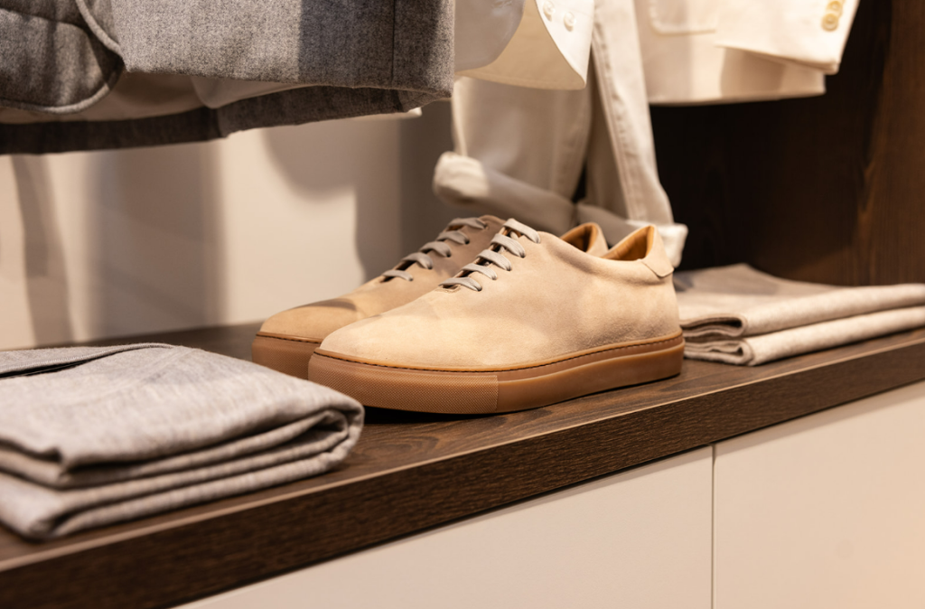 Een paar beige leren sneakers met bruine zolen staan netjes op een houten plank naast een opgevouwen lichtgrijze broek. Beige en grijze kledingstukken hangen boven en rond de schoenen, wat zorgt voor een neutrale, harmonieuze presentatie in een stijlvolle winkelomgeving. | Hoes Tailors Alkmaar