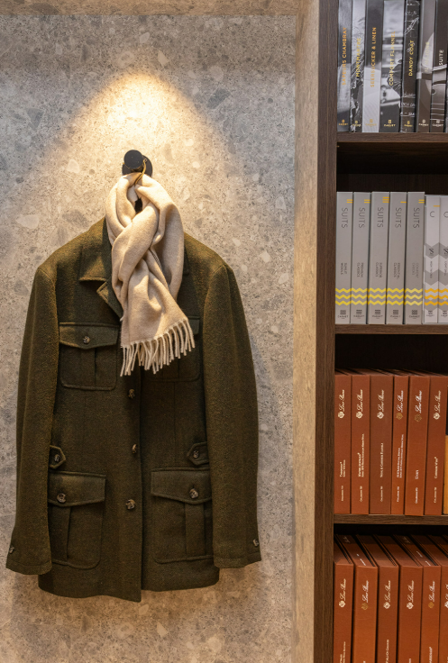 Een olijfgroene jas met grote zakken en knopen hangt aan een haakje aan de muur, met een beige wollen sjaal om de kraag. Rechts staat een boekenkast vol geordende rijen boeken en documenten in bruin, grijs en wit. | Hoes Tailors Alkmaar