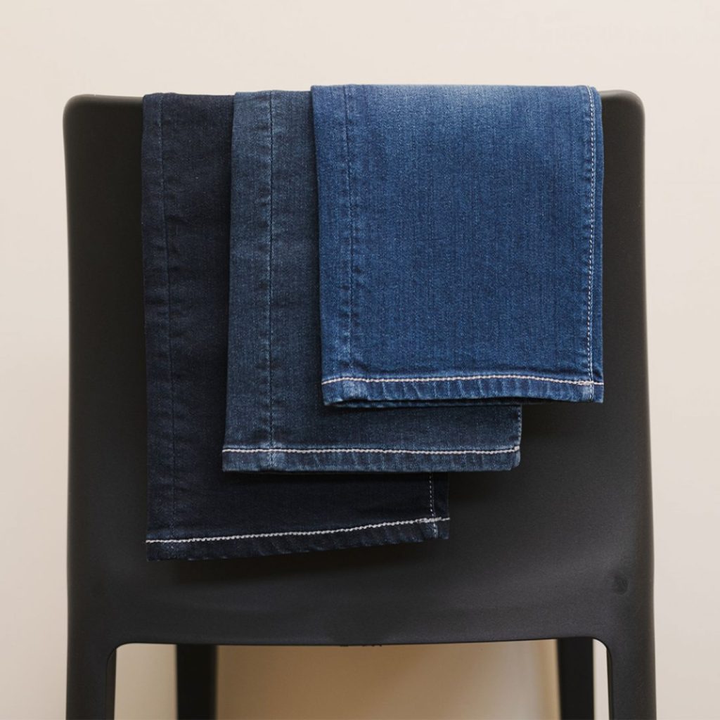 Drie spijkerbroeken in verschillende denimkleuren – zwart, donkerblauw en middenblauw – zijn netjes opgevouwen en over de rugleuning van een matte zwarte stoel gedrapeerd tegen een effen beige achtergrond. | Hoes Tailors Alkmaar